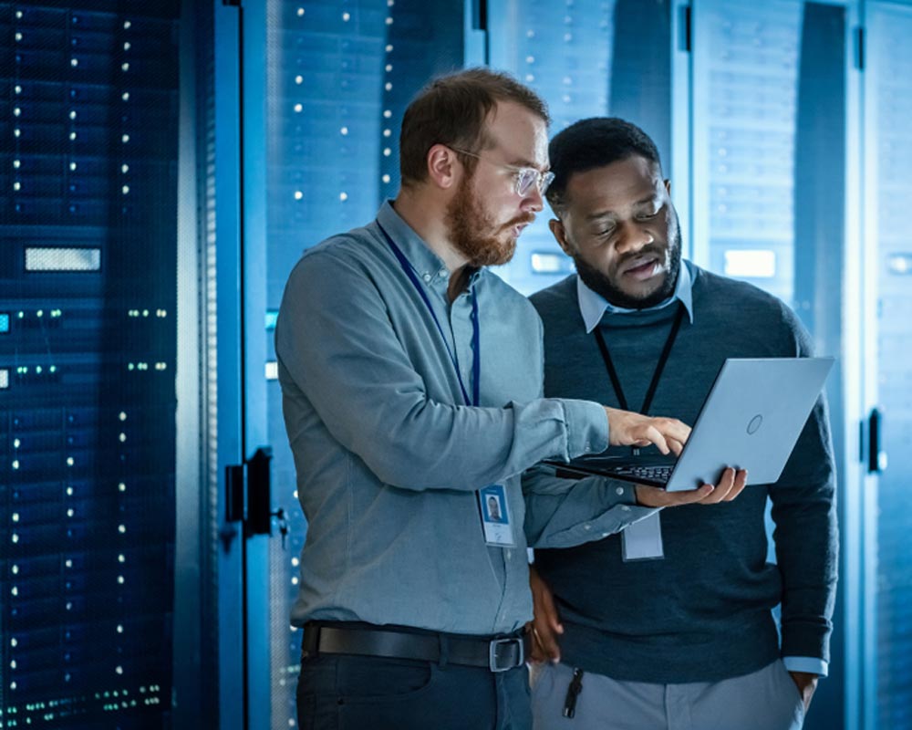 Bearded IT Technician in Glasses with Laptop Computer and Black Male Engineer Colleague are Using Laptop in Data Center