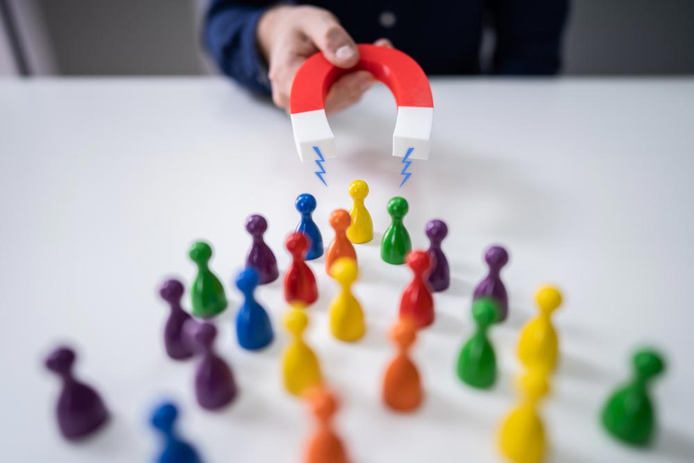 Businessperson's Hand Attracting Pawn Figures With Horseshoe Magnet On White Desk