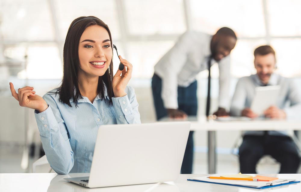 woman in headset is smiling while working with laptop in office